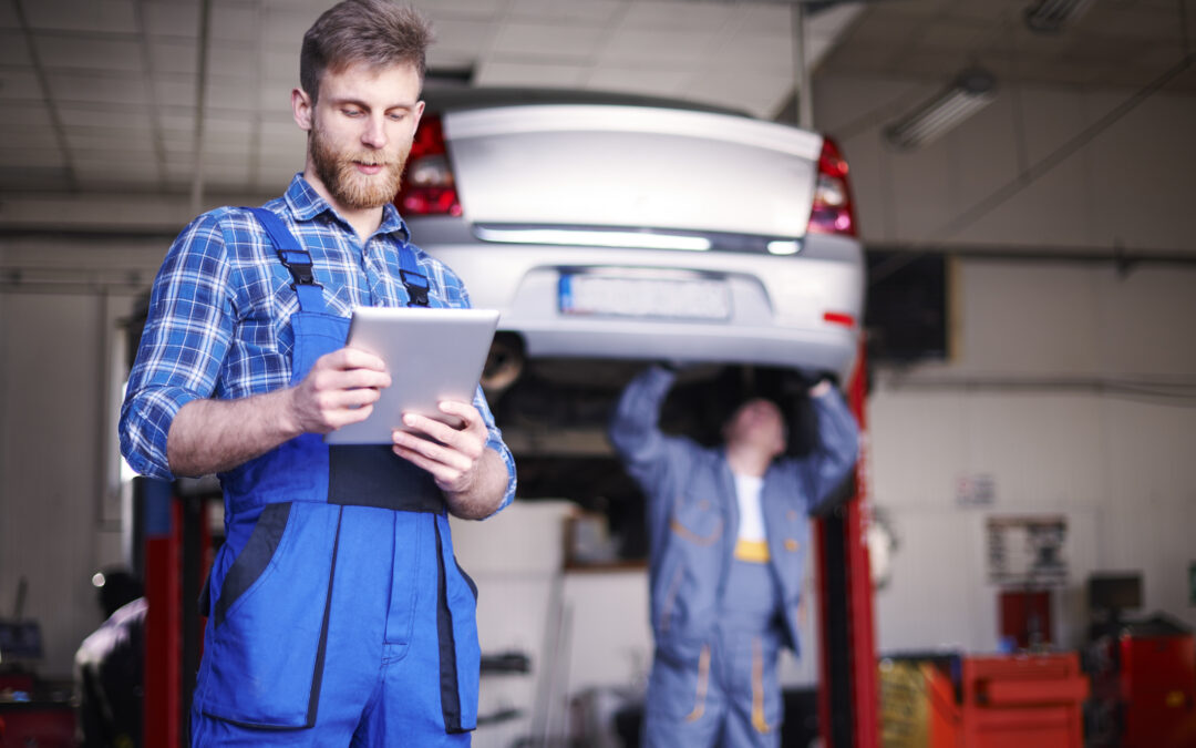Mechanic checking shop schedule using a handheld device. Another technician repairs car A/C in the background.